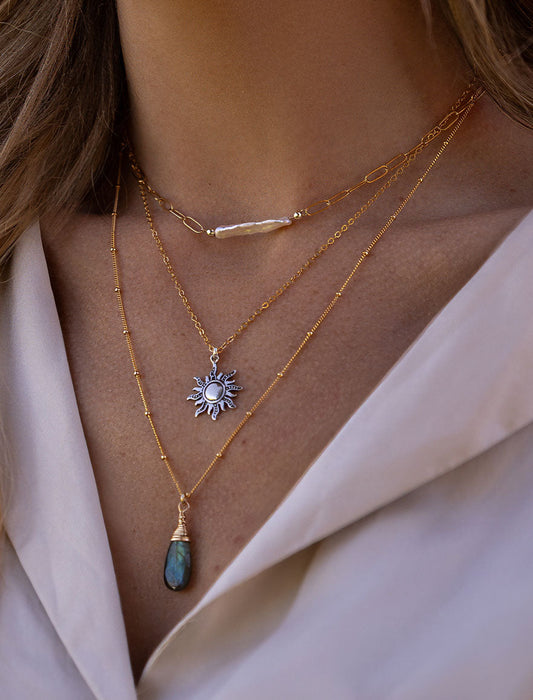 Layered gold celestial necklaces featuring mixed textures, a sun charm pendant, and a labradorite gemstone drop necklace styled on model